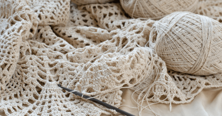 Cream-colored crocheted lace with balls of beige yarn and a crochet hook on a soft surface.