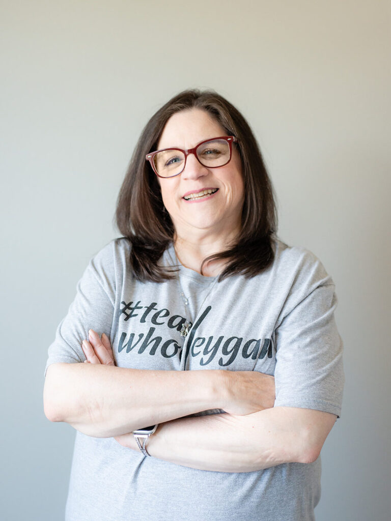 Smiling woman with red glasses and shoulder-length brown hair, arms crossed, wearing a gray T-shirt.