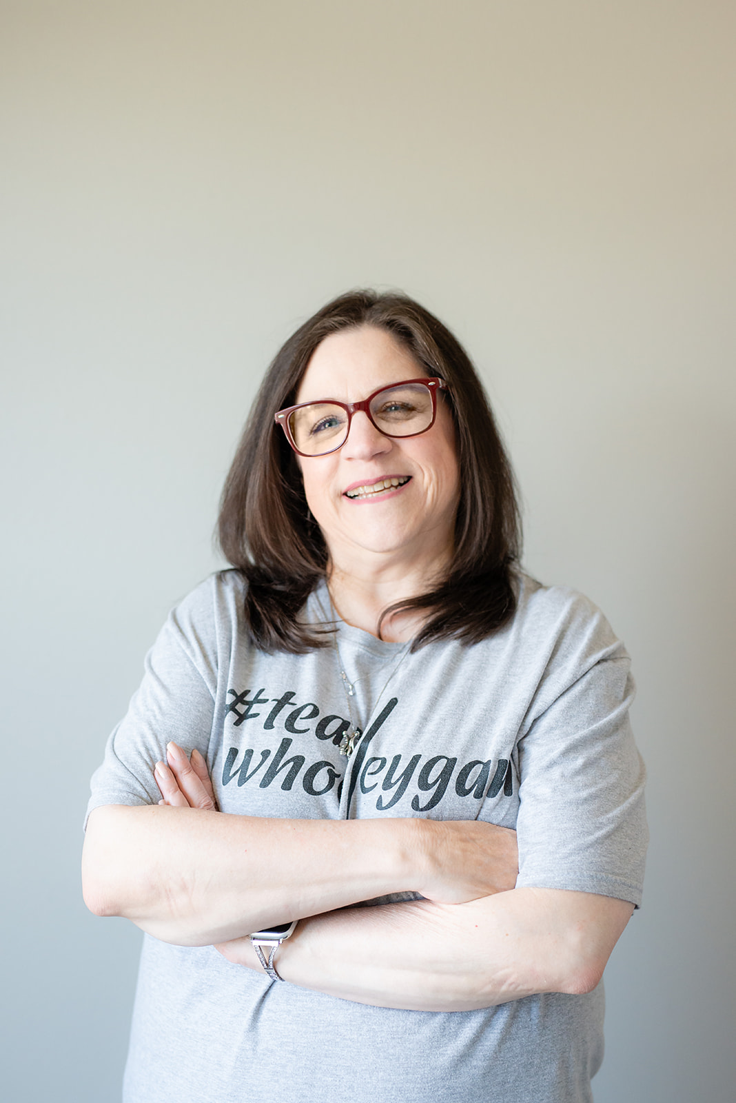 Smiling woman with red glasses and shoulder-length brown hair, arms crossed, wearing a gray T-shirt.