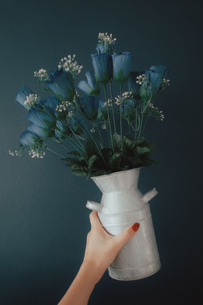 Hand holding a decorative metal vase with artificial blue rose bouquet against a dark background.