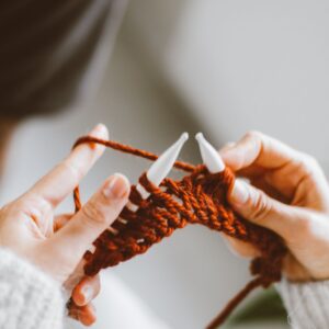 Close-up of hands knitting with orange yarn and white knitting needles.